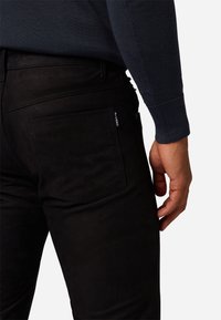 Zwarte jeans van een gladde stof, met een achterzak met een label. De pasvorm is slim met subtiele stiksels op de naden.