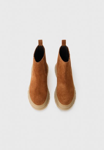 Un paio di stivali Chelsea in suede marrone con suole spesse in gomma beige, visti dall'alto su uno sfondo bianco semplice.