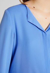 Modrá blůza s hladkým, mírně lesklým povrchem, s překlopeným límcem a jemným švem, zachycena zblízka, aby vynikly detaily.