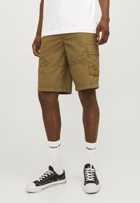 Khaki cargoshorts med flera fickor och sömdekorationer, tillverkade av lätt tyg. Kombination med svarta sneakers och vita strumpor.
