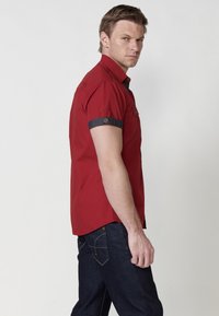 Camisa roja de manga corta con cuello abotonado, con puños enrollados en azul oscuro y detalles bordados, combinada con jeans de mezclilla oscura.