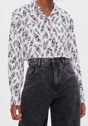 Chemise blanche courte à boutons avec des motifs abstraits noirs en spirale, portée avec un jean noir délavé taille haute sur une personne.