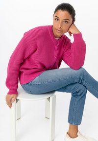Cárdigan de punto en rosa brillante con textura acanalada, con cuello redondo y tres botones, combinado con jeans azul claro y zapatillas blancas.