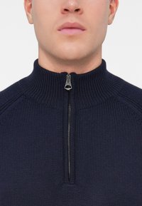 Lähivõte inimesest, kes kannab tumesinist kootud sviitrit, millel on pooleks tõmmatav kõrge krae, tõmblukk on graveeritud sõnaga "Belstaff".