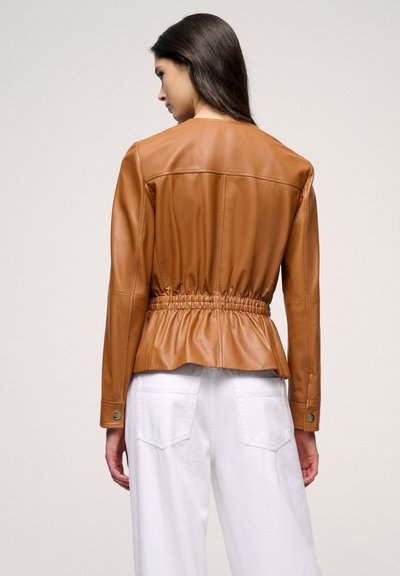 Brown leather jacket with a fitted design and a gathered waist. Features long sleeves, button cuffs, and a smooth texture. White pants visible.