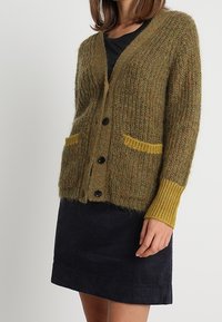 Nainen, jolla on päällään vihertävänruskea neulepusero, jossa on keltaiset taskut ja hihansuut, mustan topin päällä, yhdistettynä tummansiniseen samettihameeseen.