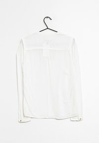 Camicia bianca a maniche lunghe appesa a un gancio nero. Tessuto leggero con polsini a bottoni, schiena plissettata e spalline sulle spalle.