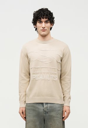 Νεαρός με σγουρά μαλλιά φοράει μπεζ φούτερ με ανάγλυφο σχέδιο του Snoopy και το κείμενο «Όλοι βιάζονται να δουν τους PEANUTS.»