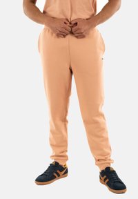 Pantalones joggers de color melocotón hechos de una suave mezcla de algodón, con bolsillos laterales y un pequeño parche con el logo. Combinados con zapatillas marinas con acentos en naranja.