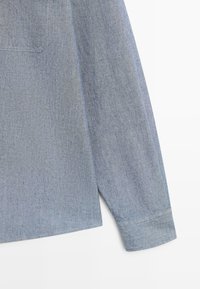 Rukáv světle modré košile z chambray s dlouhým rukávem, manžeta se zapínáním na knoflík a viditelný roh kapsy na jednobarevném bílém pozadí.