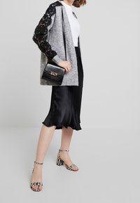 Grijze gebreide cardigan met zwarte kanten mouwen, witte top, zwarte satijnen rok en gestreepte sandalen met blokhak. Zwarte gestructureerde handtas met gouden accenten.