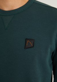 Detailní pohled na tmavě zelenou mikinu s kulatým výstřihem, na které je na hrudi vyšitý černý a oranžový logo.