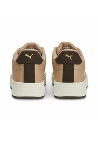 Adidași Puma cu partea superioară din material sintetic bej, accente maro și talpă albă. Dispun de un călcâi captusit și un mic logo auriu pe spate.