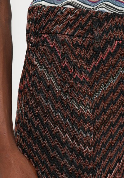 Primo piano di un braccio dalla pelle scura accanto alla vita, che indossa pantaloni a zigzag con fili neri, marroni, rosa e argento metallico.