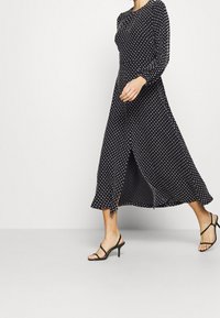 Černé midi šaty s bílými puntíky, dlouhými rukávy, nařaseným výstřihem, bočními rozparky a volnou siluetou, doplněné černými sandály na podpatku.