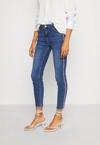 Jeans ajustados de denim azul con un acento de rayas coloridas a lo largo del costado. Presentan una cintura alta, un largo recortado y un dobladillo deshilachado.