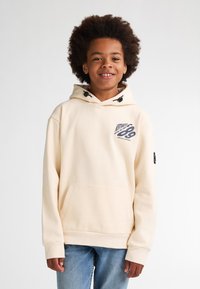 Sudadera color beige con un bolsillo delantero, capucha y cordones. Presenta un logo impreso en el pecho y una etiqueta negra en la manga. Combinada con unos vaqueros azules.