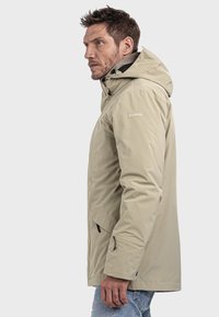 Mann in beiger Kapuzenjacke mit Schöffel-Logo auf dem Ärmel, seitlich stehend, trägt hellblaue Jeans, vor einfarbigem Hintergrund.