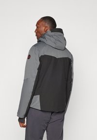 Chaqueta impermeable con la parte superior gris y la parte inferior negra, que cuenta con una capucha, bolsillos con cremallera y un parche de logo en el hombro.