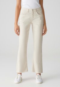 Crème-kleurige jeans met een uitlopende pijp en een ruwe zoom, met voorzakken en een ritssluiting. Gepaard met witte sneakers.