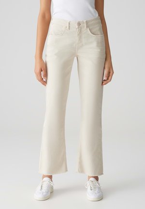 Jeans color crema con gamba svasata e orlo grezzo, dotati di tasche frontali e zip. Abbinati a sneakers bianche.