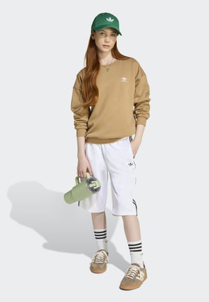 Sweatshirt beige avec logo blanc, shorts blancs avec rayures noires, casquette verte et baskets marron. Tenant une bouteille d'eau vert clair.