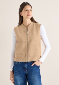 Femme portant un gilet sans manches beige zippé sur une chemise blanche à manches longues et un jean bleu, debout avec une main dans la poche, regardant de côté.