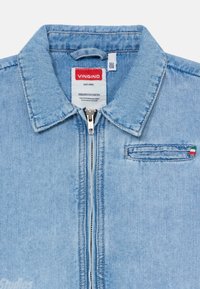 Veste en denim bleu clair avec fermeture éclair, col pointu, poche poitrine et étiquette de marque rouge. Présente un petit accent avec le drapeau italien.