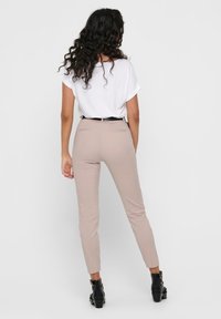 Beige nahkaleikkauksen housut, joissa on kapea malli, sileä pinta ja kaksi takataskua. Yhdistettynä löysään valkoiseen t-paitaan ja mustaan vyöhön.