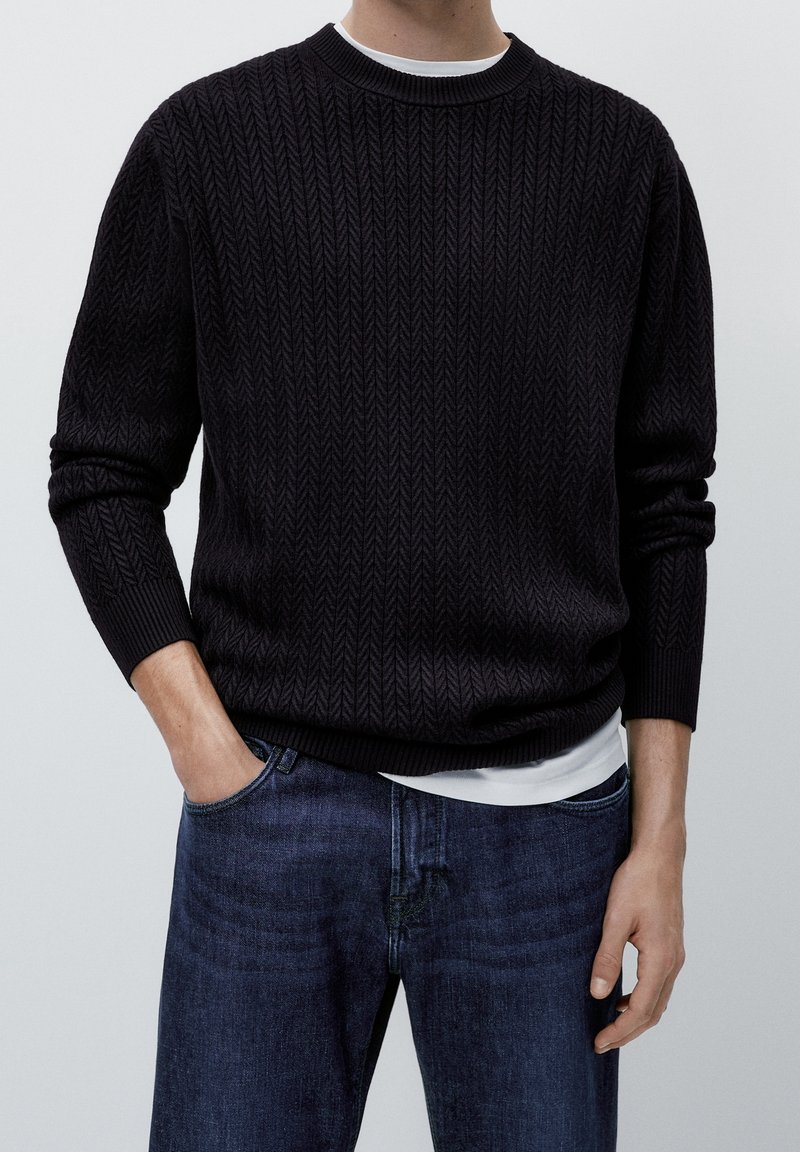 Homme portant un pull noir texturé à manches longues par-dessus une chemise blanche, une main dans la poche, jeans bleus, debout devant un fond uni.