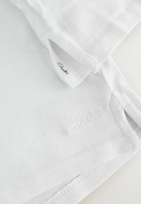 Valge T-särk lühikeste varrukatega, pehmest puuvillast materjalist, õmmeldud detailidega ja väikese tikandiga "Clarks" logoga alläär.