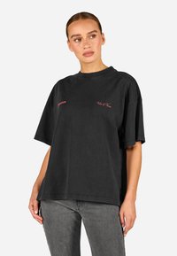 Svart oversized T-skjorte med korte ermer, laget av bomull. Har broderte røde bokstaver på forsiden. Rund hals og avslappet passform.