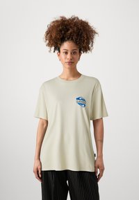 Light beige cotton t-shirt with short sleeves and a round neckline, featuring a blue graphic design on the left chest. Striped black pants.