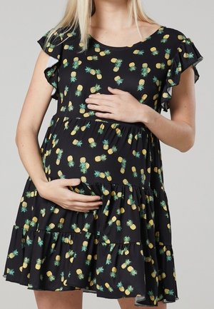 Gravid kvinde iført sort kjole med gult ananasprint, blidt krammer maven med begge hænder mod grå baggrund.