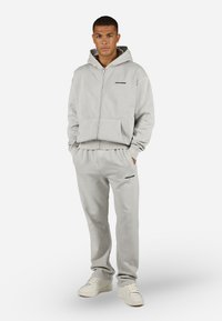 Sudadera con cremallera en gris claro y joggers a juego en material suave, con un bolsillo delantero y detalles del logotipo en negro, combinados con zapatillas blancas.