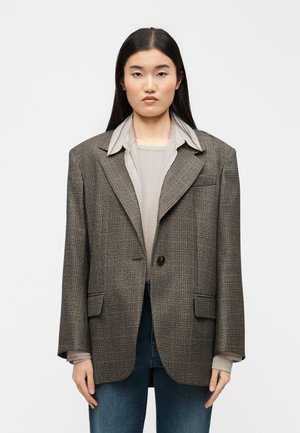 Žena s dlhými tmavými vlasmi, oblečená v nadrozmernom hnedom kockovanom saku cez svetlú košeľu a modré džínsy, stojaca pred jednoduchým pozadím.