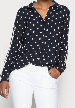 Persona che indossa una camicia a maniche lunghe blu navy con pois bianchi e strisce sulle maniche, abbinata a pantaloni bianchi, con la mano in tasca.