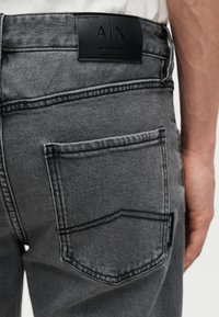 Κοντινό πλάνο πίσω τσέπης γκρι τζιν με μαύρη δερμάτινη ετικέτα Armani Exchange στη ζώνη μέσης και εμφανείς ραφές.