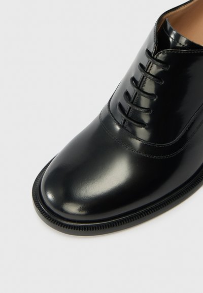 Black leather shoe with a polished finish, rounded toe, and six-eyelet lace-up design. Features subtle stitching along the upper edge and a textured sole.