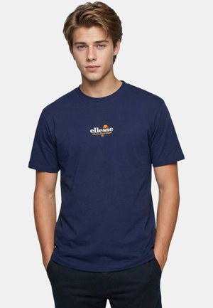 Jeune homme aux cheveux châtain clair portant un t-shirt ellesse bleu marine, debout les mains dans les poches devant un fond uni.