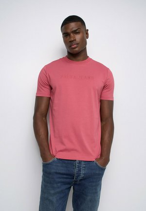Jeune homme aux cheveux courts portant un t-shirt rose uni avec l'inscription "SALSA JEANS" et un jean bleu, debout devant un fond blanc.