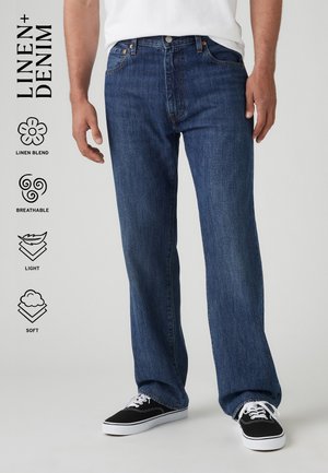 Man wearing straight-leg blue denim jeans with black and white sneakers, white shirt, and text icons highlighting linen blend, breathable, light, and soft.