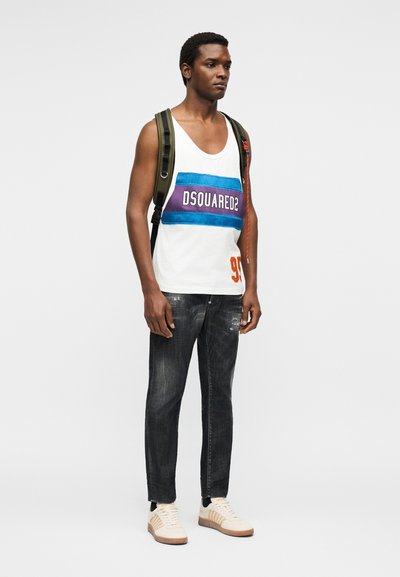 Homme portant un haut sans manches blanc avec des rayures bleues et violettes et le texte "DSQUARED2", jean noir, baskets crème et sac à dos vert.