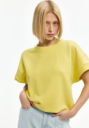 Junge Frau mit blondem Bob-Haarschnitt, trägt ein lockeres gelbes Kurzarmshirt und hellblaue Jeans mit Händen in den Taschen.