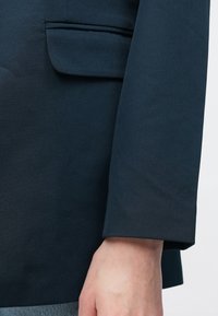 Tamsiai mėlynas blazeras su lygiu paviršiumi ir struktūrizuotu dizainu. Pasižymi viena krūtinės kišene ir sulenktais rankogaliais.