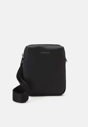 Compacte zwarte Calvin Klein schoudertas met verstelbare band en ritssluiting bovenaan tegen een effen lichte achtergrond.