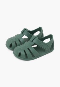 Chanclas de plástico verde con múltiples recortes, correa ajustable y plantilla texturizada. Presenta una placa con el logo en el lado.