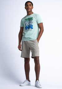 Světlězelené bavlněné tričko s víc barevným grafickým potiskem. Béžové šortky s potiskem loga. Bílé tenisky doplňují outfit.