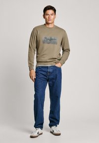 Sudadera color caqui con un gráfico oscuro que presenta "Pepe Jeans" en el frente, combinada con jeans azules y zapatillas blancas.