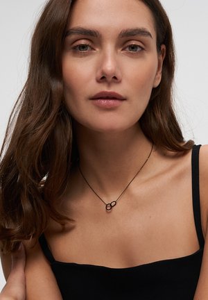 Schwarze Metallkette mit zwei ineinandergreifenden ovalen Formen, getragen auf einem nackten Schlüsselbein, vor einem einfarbigen schwarzen Oberteil. Glatte Textur, minimalistisches Design.